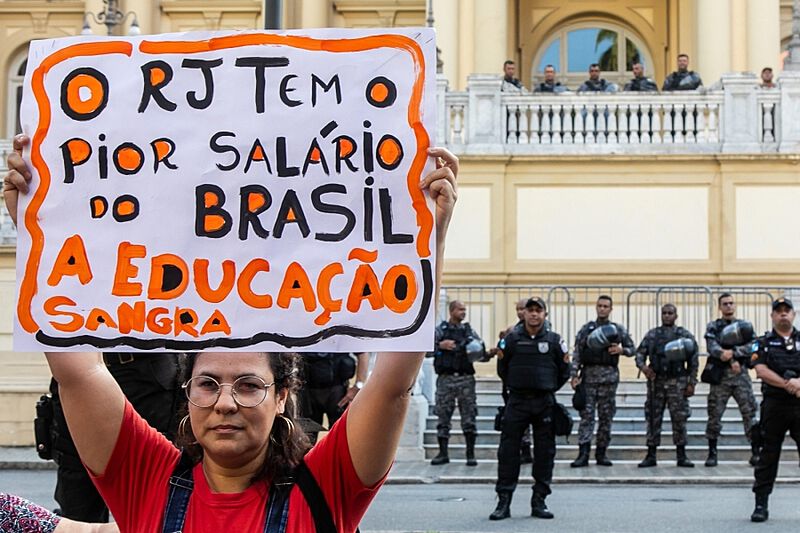 Professora em greve segura uma placa onde se lê "O RJ tem o pior salário do Brasil - a educação sangra". Ao fundo, policiais parados em frente a um prédio.
