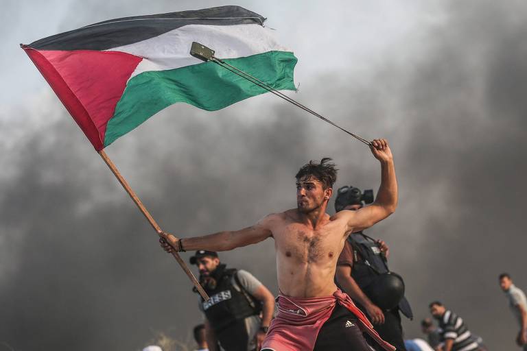 Fotografia de jovem que segura em uma mão, uma bandeira palestina erguida ao vento e, na outra, gira uma funda para lançar uma pedra.