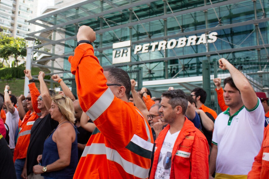 Fotografia com trabalhadores da Petrobrás, com uniformes laranja, reunidos em assembleia que votam com os punhos erguidos. Ao fundo, a fachada de um prédio da Petrobrás.