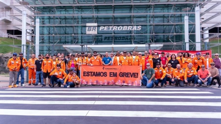 Fotografia de trabalhadores da Petrobrás com uniformes laranja reunidos em frente a um prédio da Petrobrás. Seguram uma faixa onde se lê "Estamos em greve".