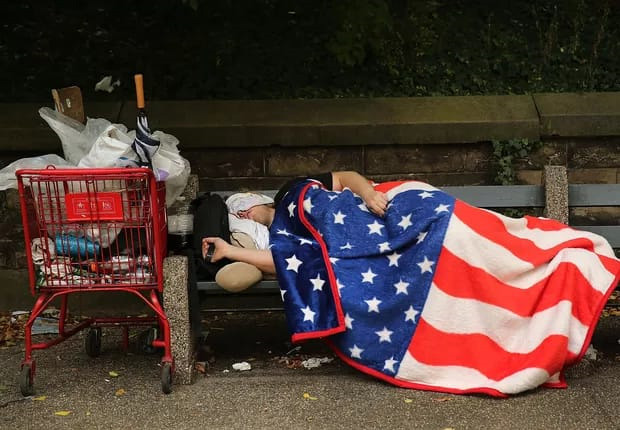 Morador em situação de rua nos Estados Unidos dorme em um banco na rua, coberto com um cobertor com o desenho da bandeira dos EUA. À esquerda, um carrinho de supermercado com os pertences do morador em situação de rua.