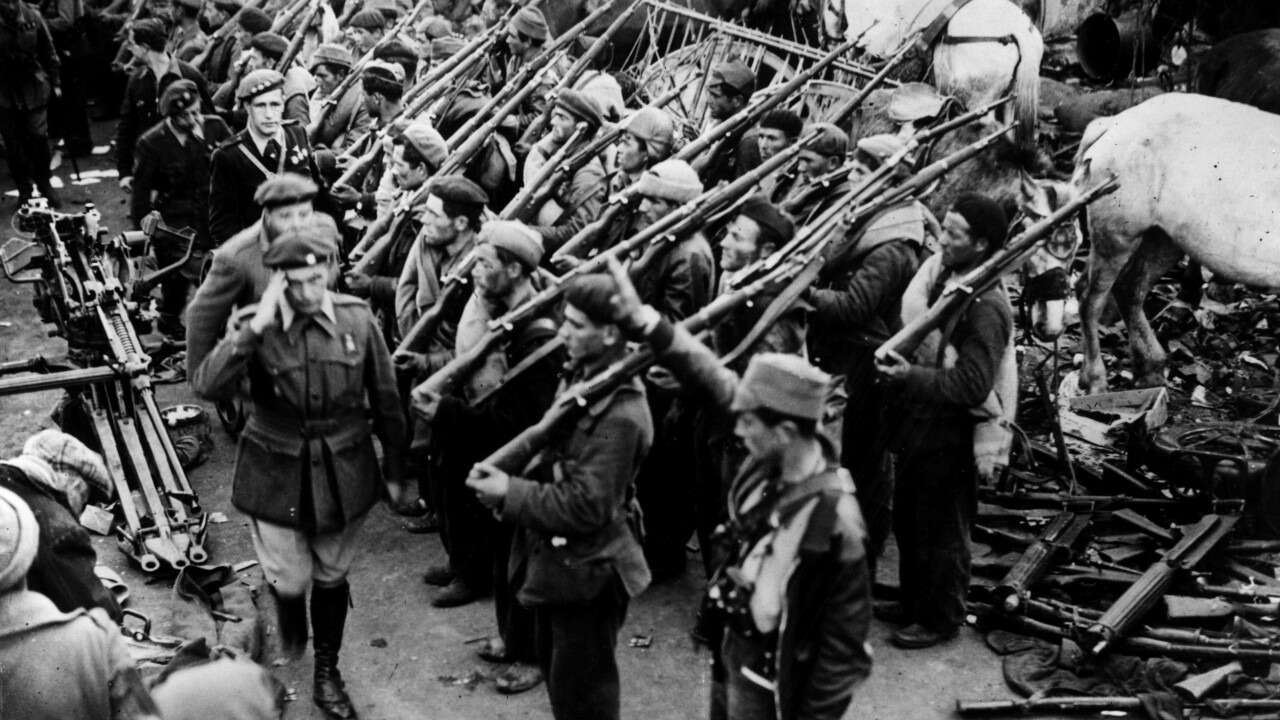 Fotografia antiga em preto-e-branco onde homens com uniforme militar passam batendo continência, enquanto outros tem carabinas e outras armas nos ombros, saudando os primeiros.