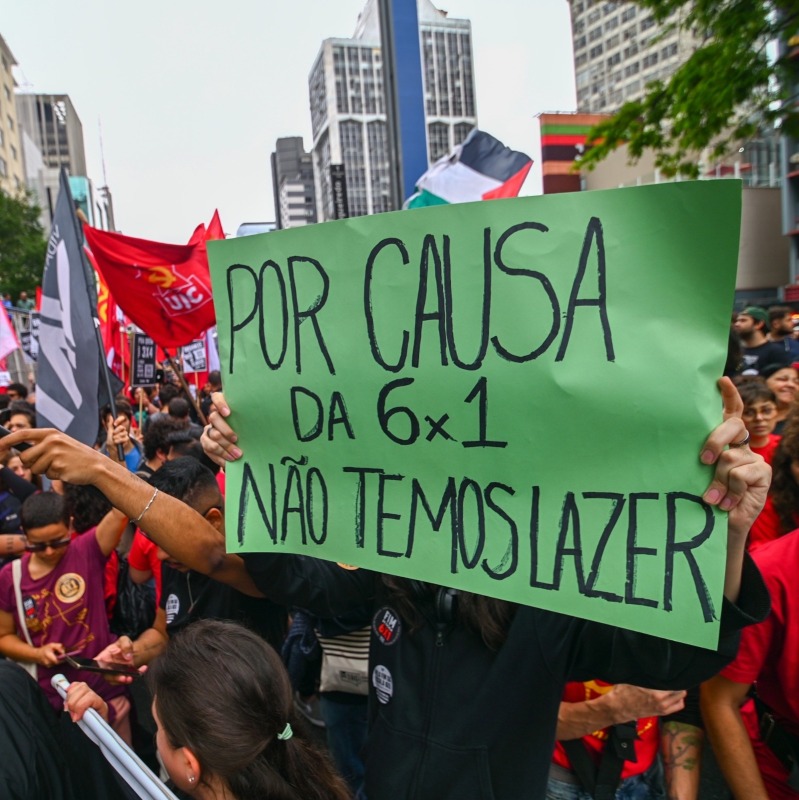Manifestação onde manifestantes seguram um cartaz verde onde está escrito "Por causa da 6x1 não temos lazer"